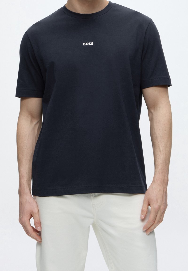 Homme portant un t-shirt noir uni à manches courtes avec le mot "BOSS" en petites lettres blanches sur la poitrine et un pantalon blanc.