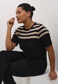 Black and beige striped knit sweater with short sleeves, round neckline, and ribbed hem, worn over black pants. Smooth texture and fitted design.