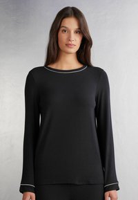Zwart langarmtop met een ronde hals en witte piping accenten bij de kraag en manchetten. Zacht materiaal, relaxte pasvorm.