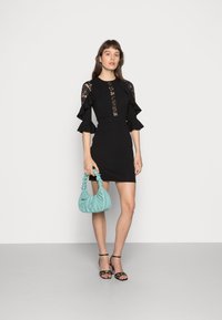 Vestido preto ajustado com detalhes em renda e mangas drapeadas, combinado com uma bolsa azul-petróleo e sandálias pretas de tiras, sobre um fundo neutro.