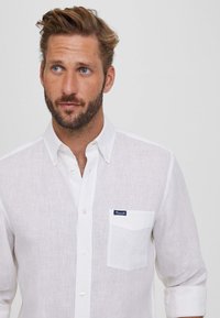 Camisa blanca de lino con cuello abotonado, bolsillo en el pecho y una textura sutil. Presenta una etiqueta de la marca en color marino en el bolsillo.