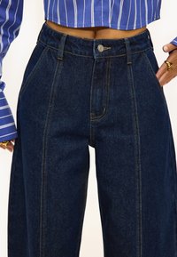 Jeans de pernera ancha en azul oscuro, hechos de mezclilla, con cintura alta, costuras amarillas y cierre frontal con botón.