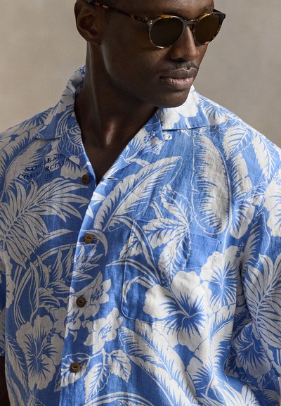 Moški, ki nosi sončna očala s okroglimi tortoisimi in modro-belo cvetlično srajco z gumbi, gleda po strani na enobarvnem ozadju.