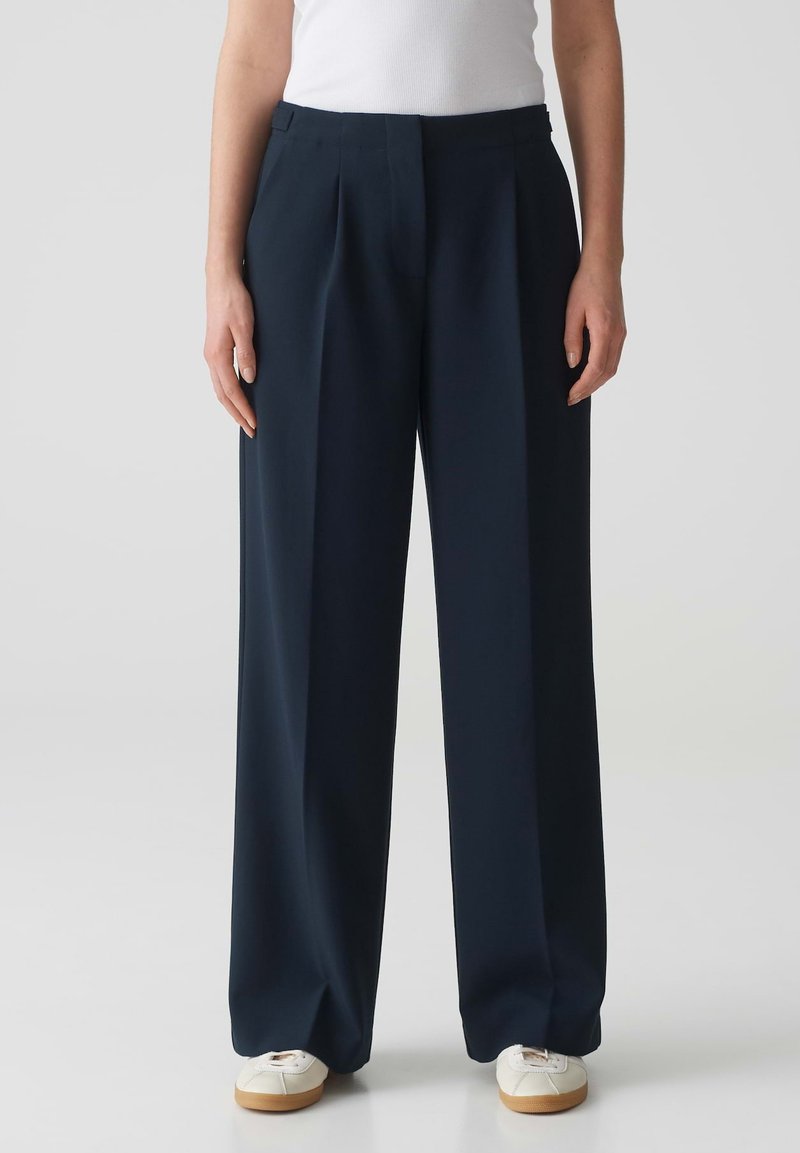 Navy wijde broek met een geplooid voorpand, op maat gemaakte snit en gladde stof. Gecombineerd met witte sneakers voor contrast.