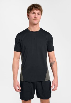 Mand med kort hår og mustache iført en sort og grå atletisk t-shirt og sorte shorts, stående mod en ensfarvet hvid baggrund.