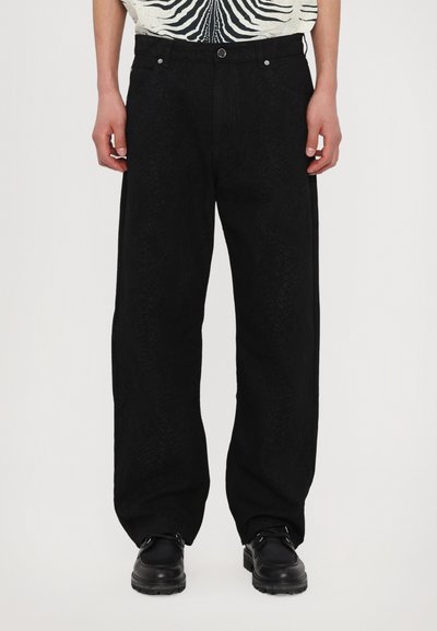 Pantalon large noir avec un motif texturé, comportant des poches avant et une fermeture à bouton. Stylisé avec une chemise à motifs et des chaussures noires.