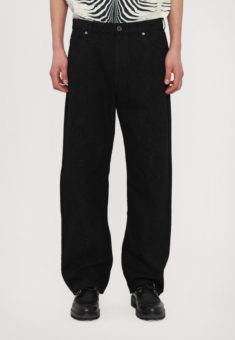 Schwarze weite Hosen mit einem strukturierten Muster, ausgestattet mit Fronttaschen und einem Knopfverschluss. Kombiniert mit einem gemusterten Hemd und schwarzen Schuhen.