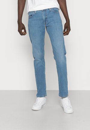 Homme portant un jean bleu clair, des baskets blanches et une chemise blanche, debout devant un fond clair uni.