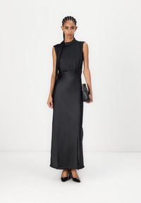 Black satin maxi dress with a high neckline, fitted waist, and side slit, paired with a small black handbag and pointed-toe heels.