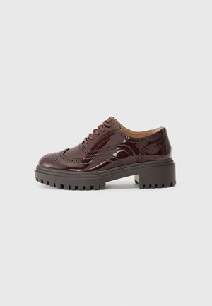 Burgundy patent leather lace-up shoe with brogue detailing, thick gray platform sole, and round toe. Textured surface and sturdy design.