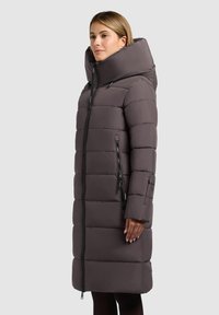 Duvet jakna u tamno sivoj boji, sa velikim kapuljačom, vertikalnim zip pukotinama i kockastim dizajnom. Glatka tekstura i dužina do koljena.