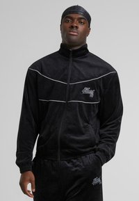 Jachetă din velur negru, cu dungi albe, guler înalt, închidere cu fermoar și accent logo. Dispune de manșete elastice și o croială lejeră.