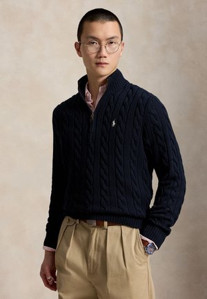 Jeune homme avec des lunettes portant un pull en maille torsadée bleu marine sur une chemise rose, un pantalon beige et une montre-bracelet, debout sur un fond neutre.