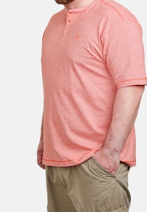 Mężczyzna w koralowej koszulce henley z krótkim rękawem i trzema guzikami oraz khaki szortach typu cargo, z ręką w kieszeni, stoi bokiem.