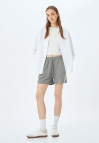 Witte button-up overhemd, aansluitend wit crop-top, grijze shorts met trekkoord en logo, witte sneakers met grijze en bruine accenten, en witte sokken.