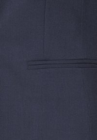 Tissu texturé bleu marine avec un motif en grille subtil, doté d'une poche passepoilée et de coutures nettes et précises pour un look sur mesure.