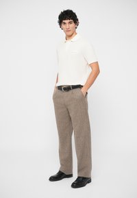 Νεαρός άνδρας με σγουρά μαλλιά, φορώντας λευκό πόλο με κοντά μανίκια, καφέ υφασμάτινο παντελόνι με μαύρη ζώνη και μαύρα κοστούμια παπούτσια, που στέκεται μπροστά σε λευκό φόντο.