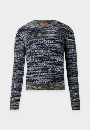 Kvinde iført en mørk mønstret sweater, vidtbenede blå jeans og åbne hæle, stående med den ene hånd bag ryggen på en ensfarvet hvid baggrund.
