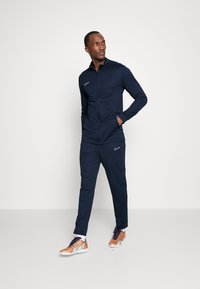 Marineblauwe sporttracksuit met een ritsjack en broek, voorzien van een wit Nike-logo. Sneakers in koper met witte accenten. Soepele stof tekstuur.