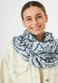 Écharpe légère à motifs dans des tons de bleu et de blanc, avec une finition douce et texturée, dotée de bords effilochés, enroulée autour du cou.