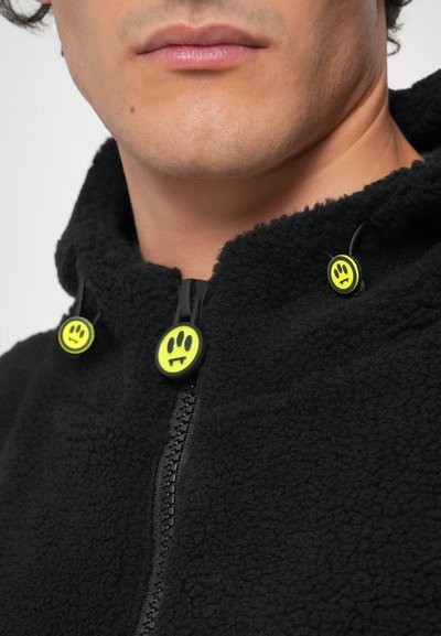 Fekete bolyhos dzseki cipzáros záródással, sárga mosolygós arccal díszített zsinórokkal és strukturált felülettel. Kapucnis kialakítás, zsinóros állíthatósággal.