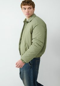 Jasnozielona kurtka puchowa z kołnierzem, przednim zamkiem błyskawicznym i elastycznymi mankietami. W zestawieniu z ciemnoniebieskimi dżinsami z tylną kieszenią.