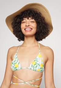 Biquíni triangular em branco com estampado de limões amarelos e folhas verdes; possui alças finas e um design cruzado nas costas. Usado com um chapéu de palha de aba larga.
