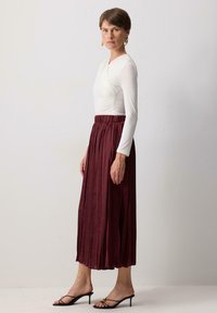 Haut à manches longues blanc et jupe maxi plissée bordeaux avec une taille élastique. Des sandales à talons noires complètent la tenue.