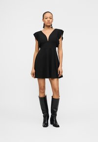 Vrouw staand, gekleed in een zwarte korte jurk met gerimpelde mouwen en kniehoge zwarte laarzen, tegen een effen lichte achtergrond.