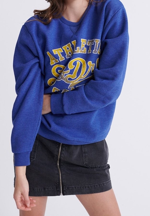 Personne portant un sweat-shirt bleu "Athletic S. Dr" et une jupe en jean noire avec fermeture éclair à l'avant, debout avec un bras plié devant le corps.
