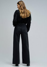 Top negro, transparente, con volantes y mangas largas, combinado con pantalones negros de tiro alto y pernera ancha. Se aprecian detalles de tejido texturizado y costuras sutiles.