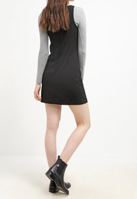 Kvinna iklädd en svart ärmlös klänning över en grå långärmad tröja, matchad med svarta ankelstövlar, stående mot en vit bakgrund.