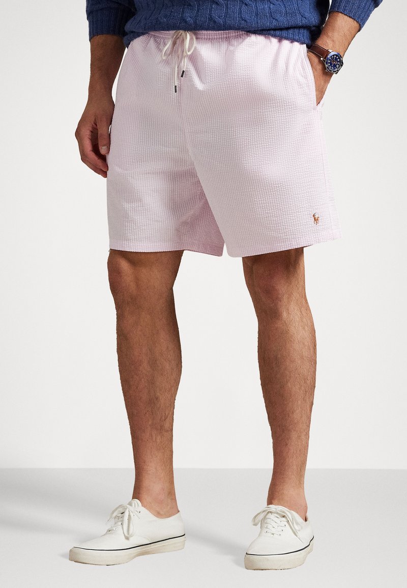 Šviesiai rožiniai maudymosi šortai su tekstūruotu audiniu, elastinga juosta, raišteliu ir mažo logo detale šalia krašto. Dėvimi su baltais sportbačiais.