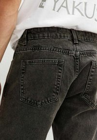 Primo piano di un paio di jeans in denim nero, che mostra il retro con due tasche posteriori e la cintura, indossati da una persona in una maglietta bianca.