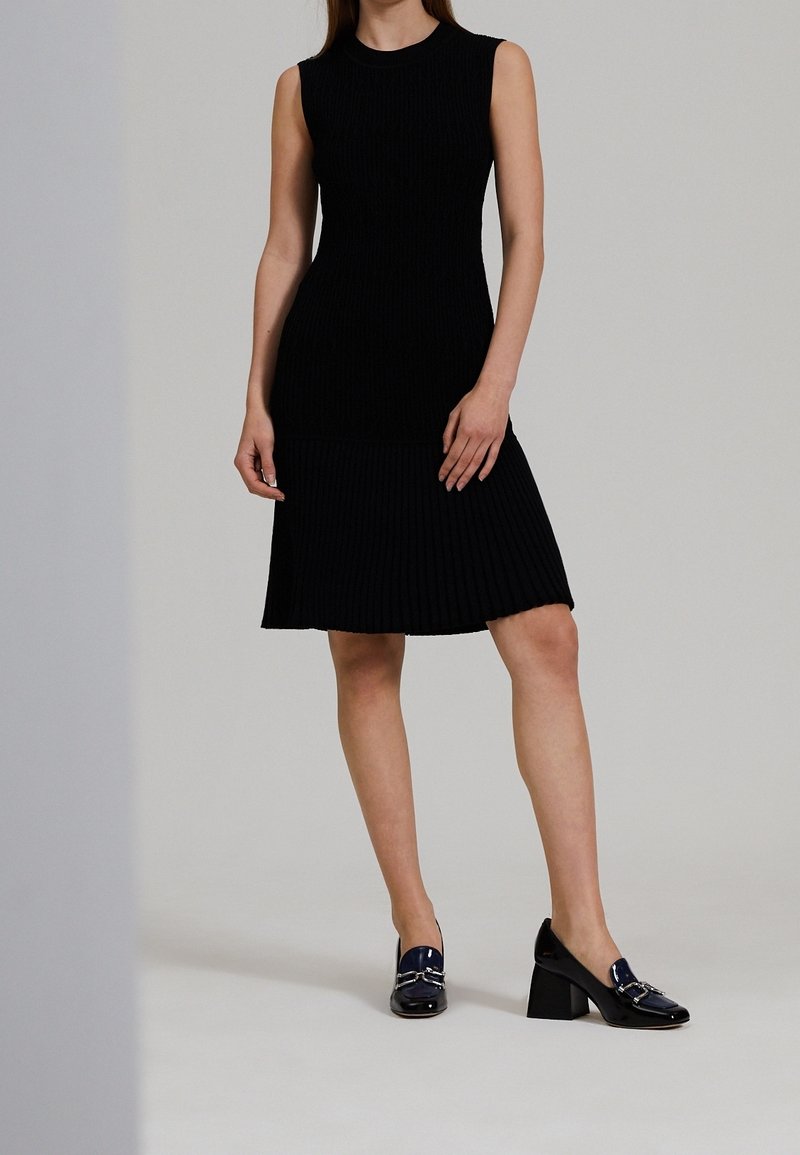 Femme portant une robe noire plissée sans manches associée à des mocassins à talons carrés bleu foncé, debout devant un fond uni.