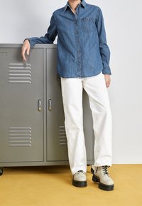 Jeansskjorta med en ficka, matchad med vita vida byxor och beiga snörade stövlar med svarta sulor, framför ett grått skåp.