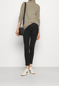 Person med en beige kabelstickad polotröja, svarta smala byxor, vita sneakers med svarta ränder och bärande en liten svart axelväska.