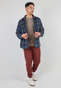 Chemise en flanelle à carreaux bleus et verts, pull beige en dessous, joggings bordeaux, baskets blanches, et matériaux en mélange de laine texturée.