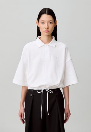 Polo bianca a maniche corte con patta con bottoni e coulisse in vita, indossata sopra pantaloni neri a vita alta con dettagli a bottoni.