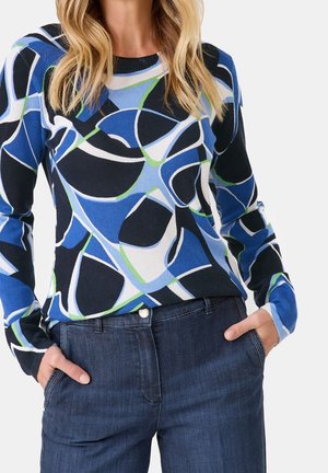 Knit sweater with a bold abstract pattern in navy, blue, green, and white. Long sleeves and a round neckline. Worn with dark denim jeans.