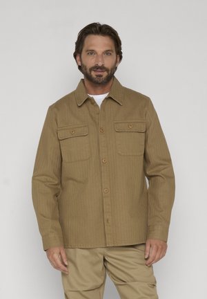 Mężczyzna o średniej długości brązowych włosach i brodzie, ubrany w karmelową koszulę na guziki z kieszeniami na piersi oraz khaki spodnie, stojący na jednolitym tle.