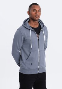 Hoodie gris zippé avec une capuche à cordon, des poches frontales et un matériel blanc contrasté, associé à un t-shirt graphique noir en dessous.