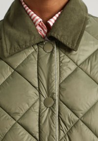 Chaqueta acolchada de color verde oliva con cuello de pana y cierre de botones, con un patrón de diamantes y ribetes en contraste a lo largo de los bordes.