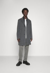 Cappotto grigio in lana con colletto strutturato, tre bottoni anteriori e tasche laterali, abbinato a pantaloni grigi chiari e scarpe nere.