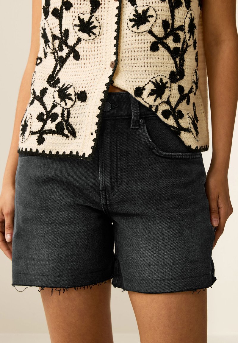 Svarta denimshorts med fransad nederkant, matchade med en krämfärgad stickad väst med svart blommönster och knäppning.