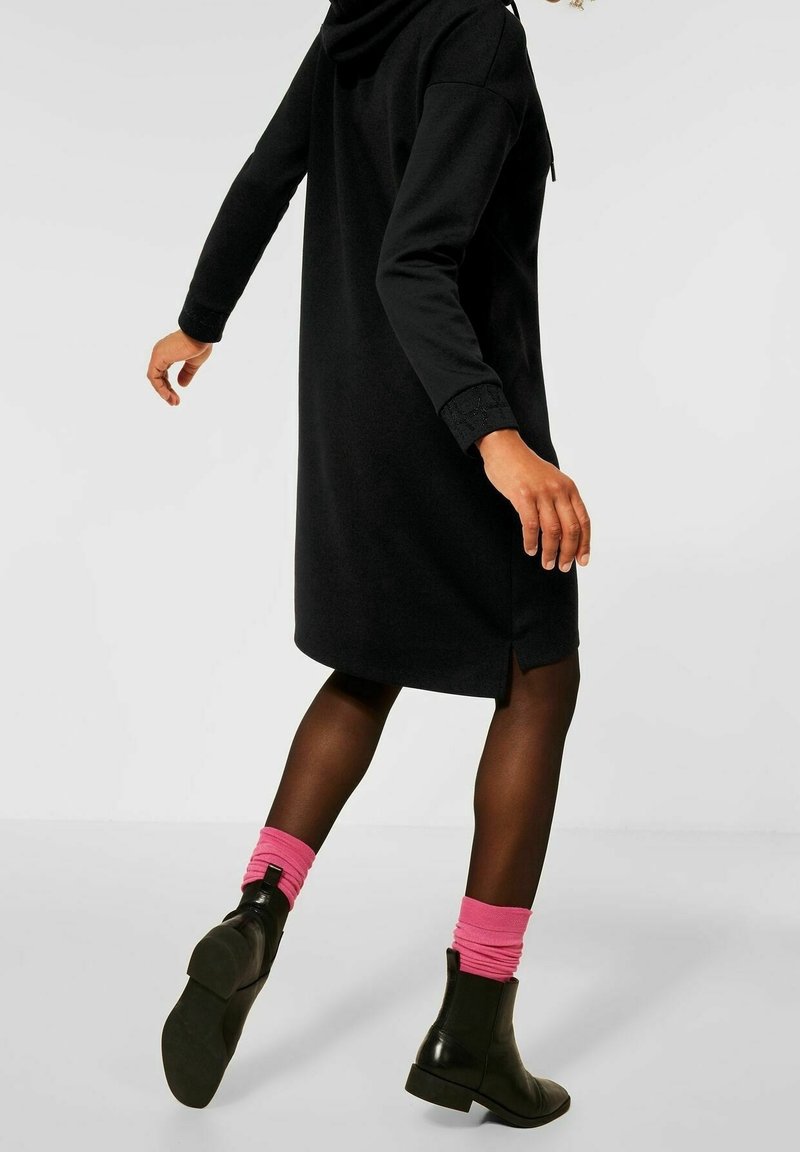 Svart huva klänning med långa ärmar, med sidofickor. Bärs med svarta ankelstövlar och rosa uppvikta strumpor, vilket framhäver texturkontrasten.