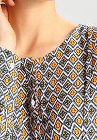 Blusa a fantasia con un motivo a diamante nei colori blu, giallo e rosso, completata da bottoni bianchi. Il tessuto ha una consistenza liscia.