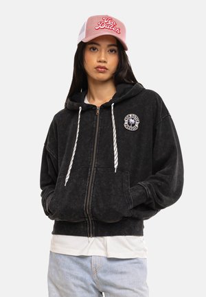 Jeune femme portant une casquette rose Von Dutch, un sweat à capuche noir zippé avec des cordons blancs, et un jean bleu clair, debout les mains dans les poches.