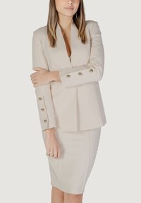Blazer beige su misura con bottoni dorati sulle maniche, abbinato a una gonna a tubino attillata dello stesso colore. Texture del tessuto liscia.