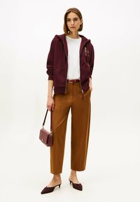 Bordeaux zippered hoodie over en hvid t-shirt, parret med brune vide bukser, bordeaux bælte, taske og sko. Glatte teksturer og afslappet stil.